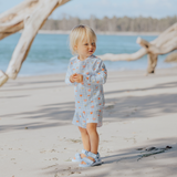 Child in a blue checkered swim set standing on a sandy beach with driftwood and water in the background.