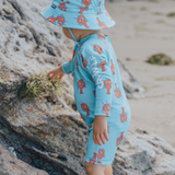 Child wearing a blue swimsuit and swimhat with orange lobster patterns on a rocky beach.
