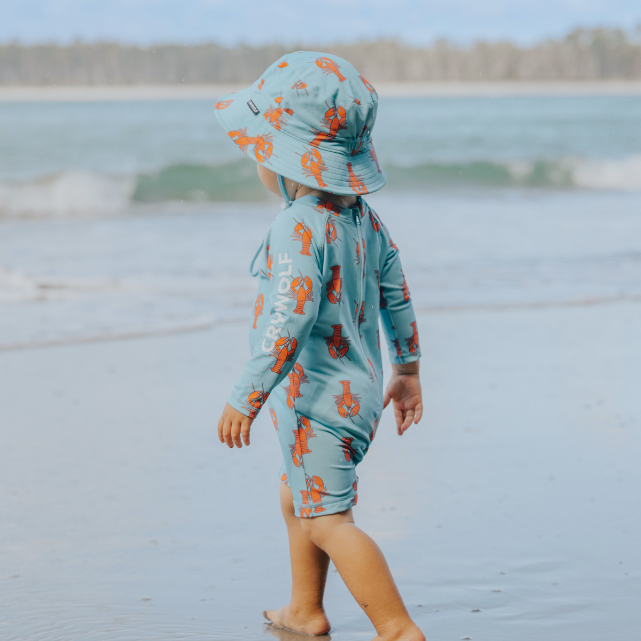 Child wearing a blue swimsuit with orange patterns and a matching hat, standing on a beach.