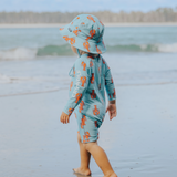 Child wearing a blue swimsuit with orange lobster patterns and matching hat on a beach.