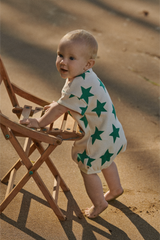 Baby in a star-patterned outfit standing on sand near a wooden chair.