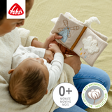 Baby holding a soft book with a windmill design, sitting on a person's lap, with Fehn logo and organic cotton certification.