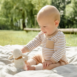 Baby sitting outdoors on a blanket with a soft toy, surrounded by greenery.