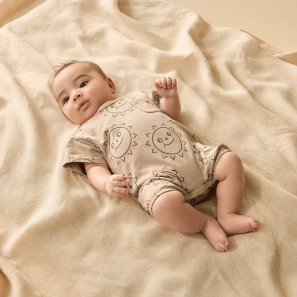 Baby lying on a beige blanket wearing a onesie with sun designs.