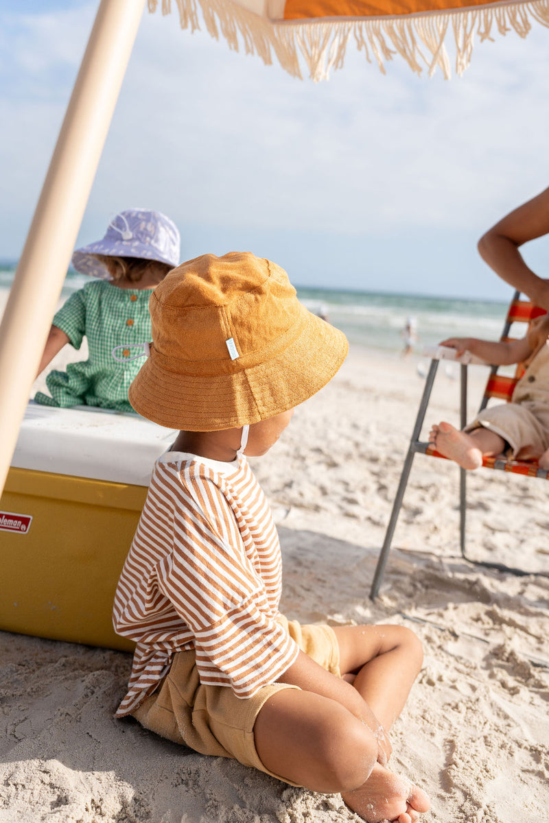 TODDLER SITTING ON SAND WEARING ACORN Terry Towelling Bucket Hat - Caramel SIDE VIEW