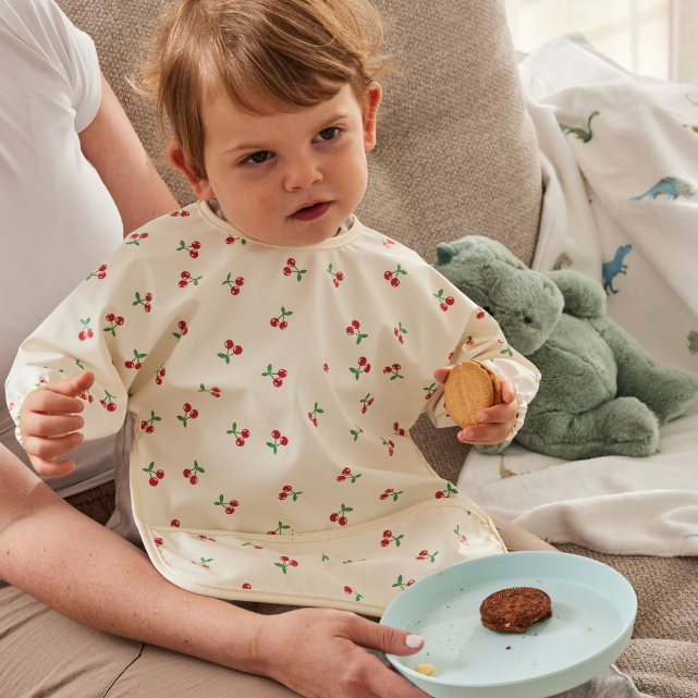 Child wearing a bib with a cherry pattern, holding a cookie, sitting on a couch with a green stuffed dinosaur toy.