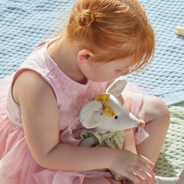 Young girl in a pink dress holding a white plush toy with a yellow bow on a textured blue and green surface.