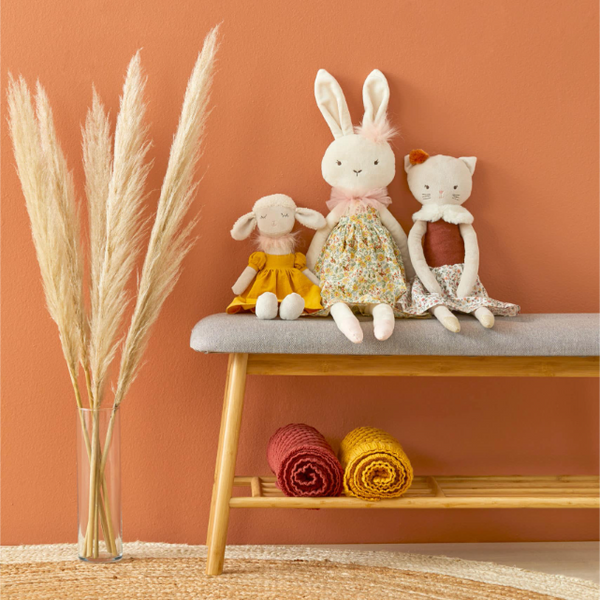 Three stuffed animals on a small table with a vase of dried plants against an orange wall.