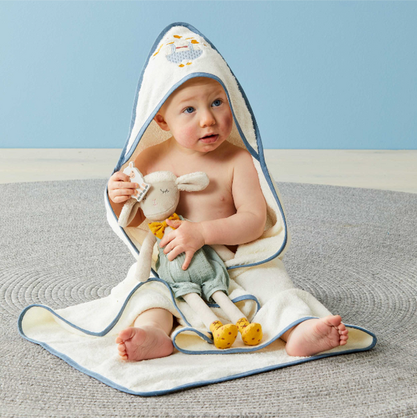 Baby wrapped in a hooded towel with a toy sheep, sitting on a textured surface with a light blue background.