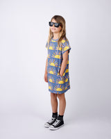 Child wearing a blue dress with yellow sun patterns and black sunglasses on a white background