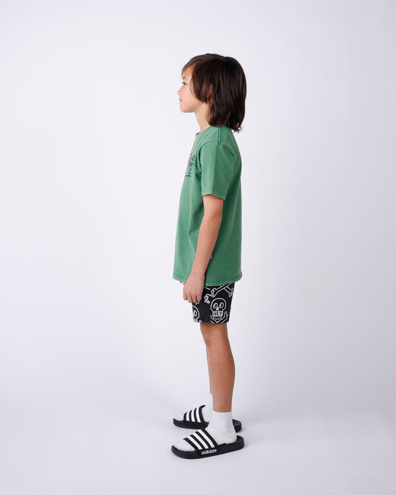 Child wearing a green t-shirt and black shorts with white patterns on a white background