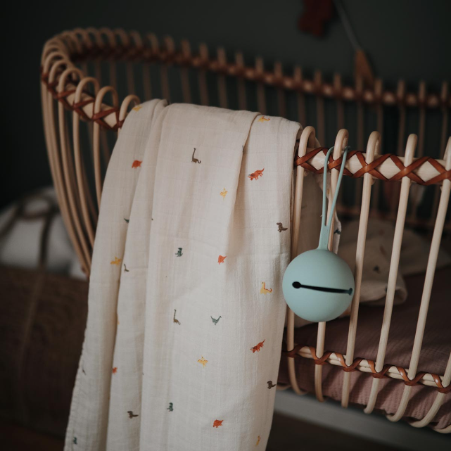 Baby crib with a soft dinosaur muslin blanket and a pacifier holder in a warm, dimly lit room.