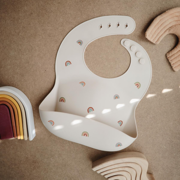 Beige bib with rainbow pattern on a brown surface with toys around