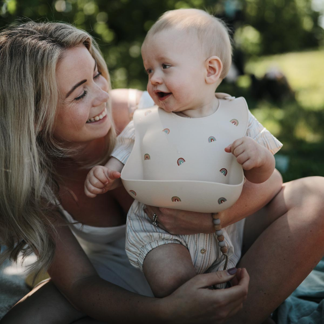 Woman holding a baby outdoors with a blurred green background. Baby wearing silicone rainbow bib