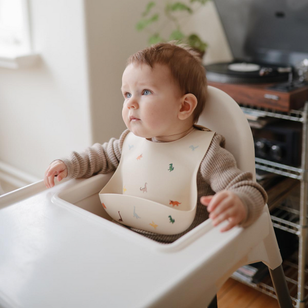 Baby sitting in a high chair wearing a silicone dinosaur bib with a neutral background