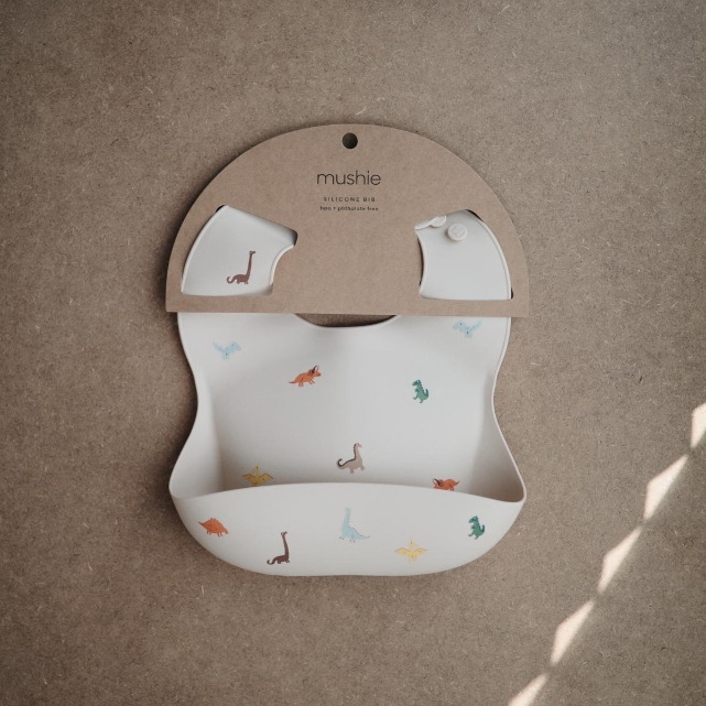 Beige Baby bib with dinosaur print on a cardboard backing, brand 'mushie' visible