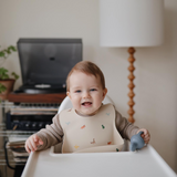 Baby sitting in a high chair with a bib and toy, smiling indoors.