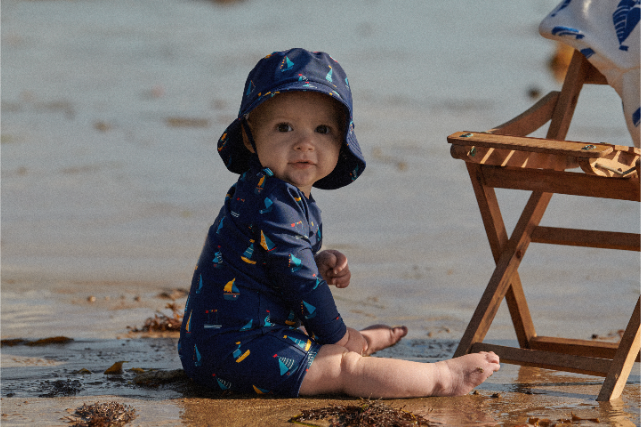 Baby in a blue swimsuit and hat sitting in shallow water at the beach.