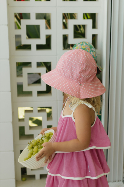 Child wearing a pink dress and hat holding a basket of grapes, standing in front of a decorative white screen.
