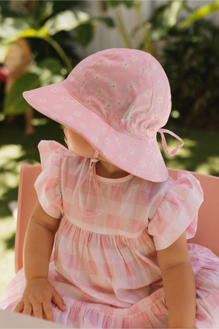 Side view of child wearing a pink dress and pink sun hat with daisy pattern, sitting outdoors with greenery in the background