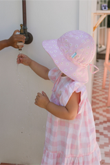 Child in a pink dress and sun hat playing with water from a faucet.