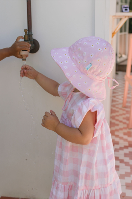 Child in a pink dress and sun hat playing with water from a faucet.