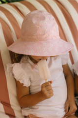 Child wearing a pink sun hat with floral patterns, holding a popsicle against a striped background.