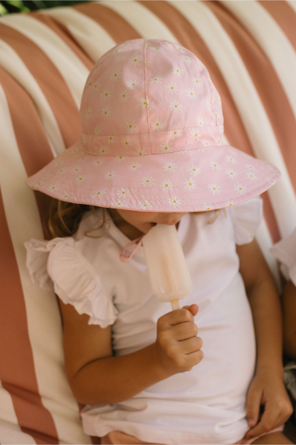 Child wearing a pink sun hat with floral patterns, holding a popsicle against a striped background.