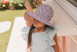 Girl wearing a floral sun hat and light blue dress under an umbrella outdoors.