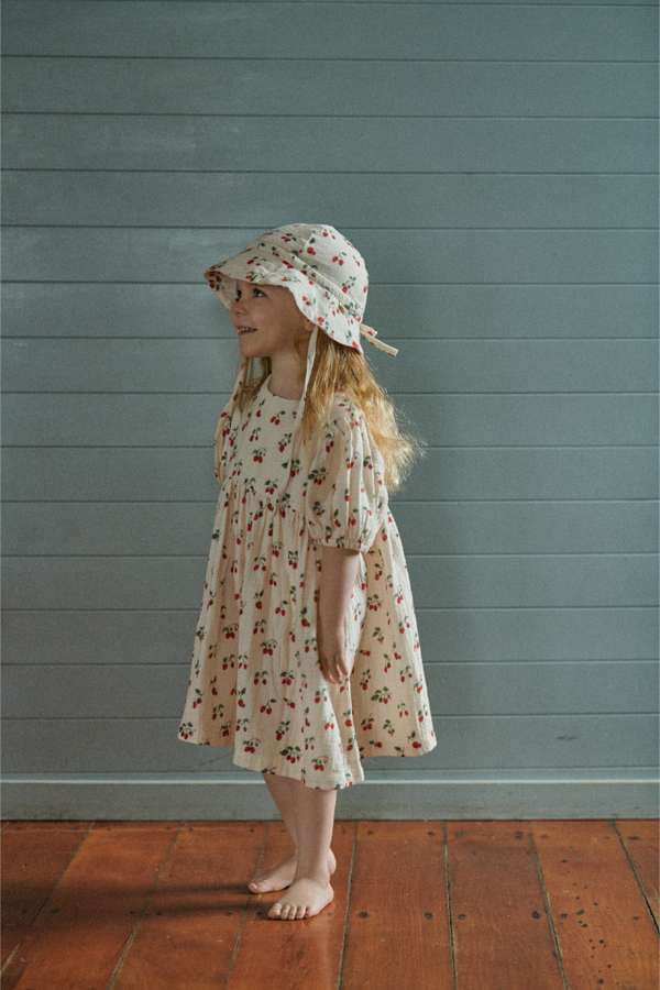 Young girl in a strawberry dress and matching hat standing against a teal wall.