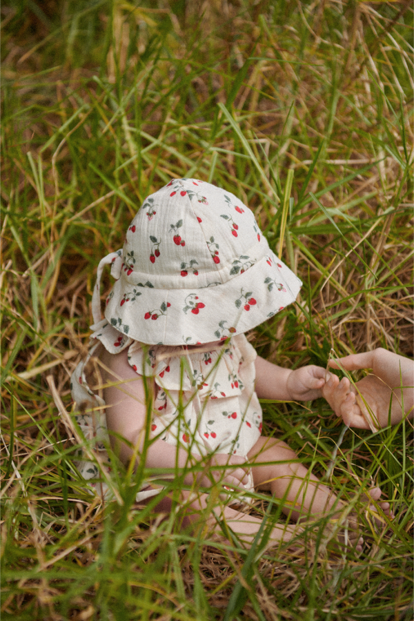 Baby in a strawberry patterned sun hat and matching romper sitting in grass 