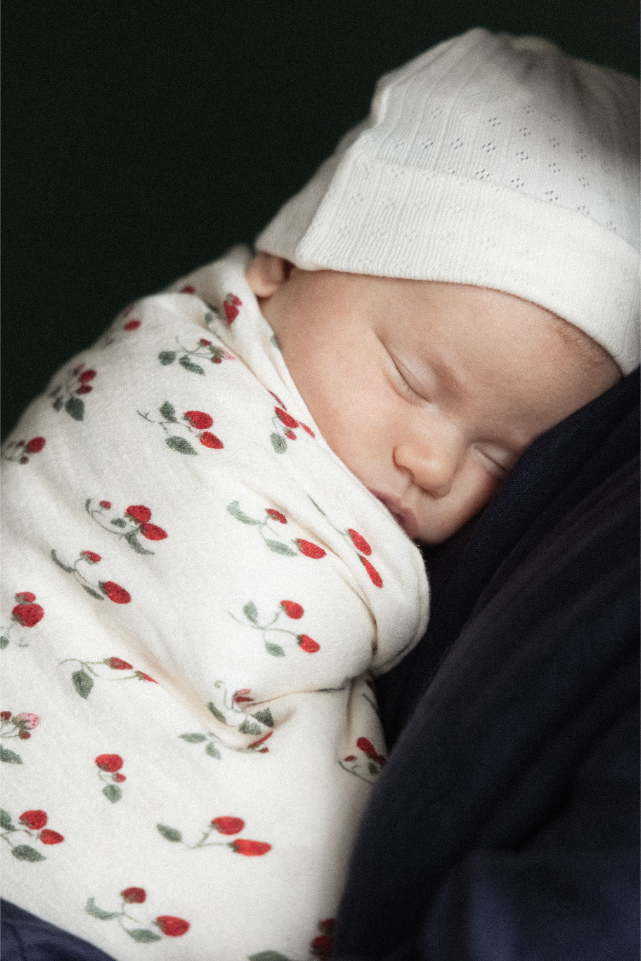 Newborn baby swaddled in a strawberry patterned blanket with a white hat on a dark background