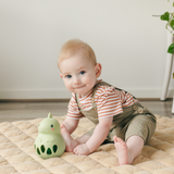 Baby sitting on a quilt with a green dinosaur toy, wearing a striped shirt and overalls.