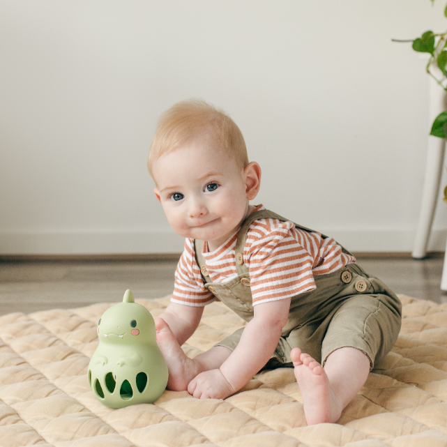Baby sitting on a quilt with a green dinosaur toy, wearing a striped shirt and overalls.