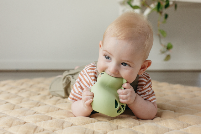 Baby chewing a green teething toy on a beige blanket