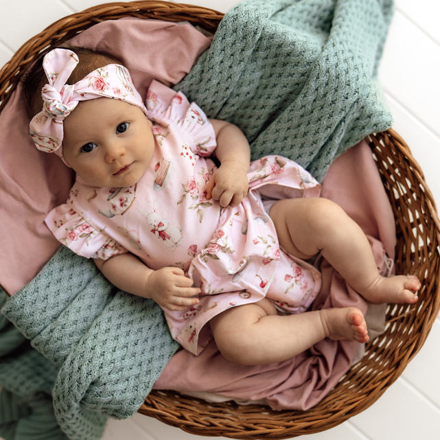 Baby in a floral outfit lying in a wicker basket with pink and green blankets.