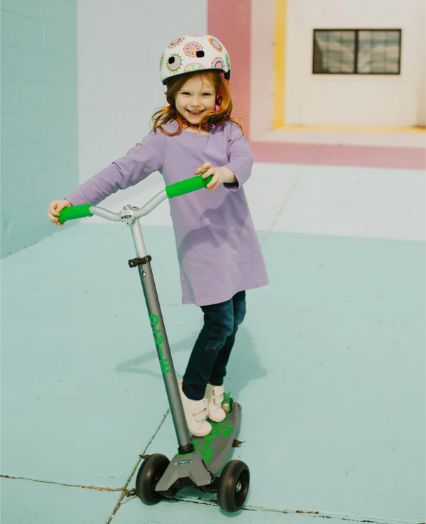 Child in a purple dress and helmet standing on a scooter in a colourful room.