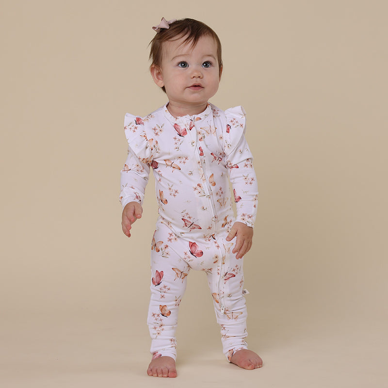 Baby standing wearing the SNUGGLE HUNNY Butterfly Organic Snugglesuit Convertible Romper with Frill